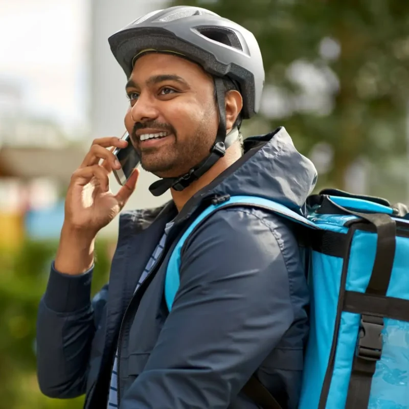 Homem com capacete e mochila azul falando ao celular ao ar livre, em ambiente urbano, sugerindo contratação de seguro acidente durante atividades diárias