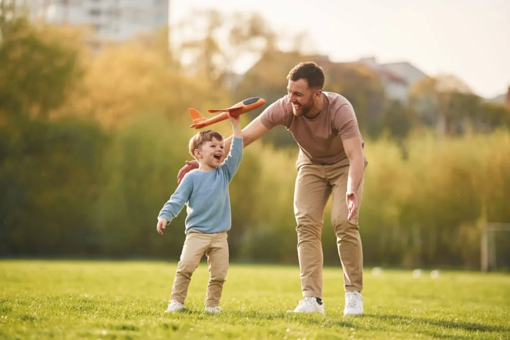 svg+xml;charset=utf em Maximídia - Seguro de Acidentes Pessoais e Seguro de Vida com o Máximo de Benefícios Pai brincando no parque com uma criança em um dia ensolarado, segurando o brinquedo e incentivando a diversão ao ar livre — seguro contra acidentes pessoais